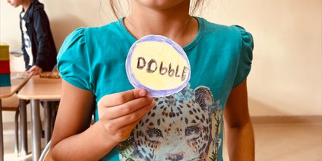 Powiększ grafikę: Na zdjęciu dziewczynka stoi w niebieskiej koszulce z okrągłym Dobble, w tle ściana.a