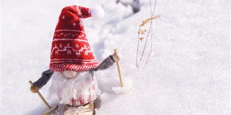 Powiększ grafikę: Grafiki poglądowa - kukiełka świątecznego elfa z czerwoną czapką i brodą na nartach, pośród śniegu.