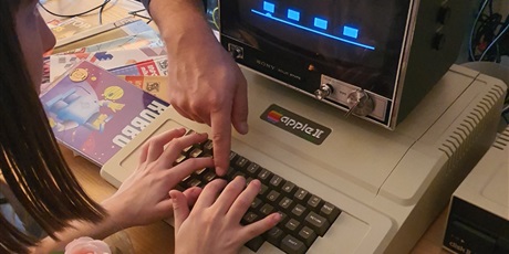 Powiększ grafikę: Opiekun zajęć wskazuje uczennicy, co ma wcisnąć na klawiaturze starego komputera.