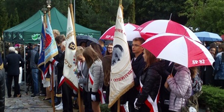 Powiększ grafikę: Uczniowie z pocztu sztandarowego SP82 stoją pod parasolami na cmentarzu.