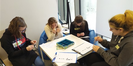 Powiększ grafikę: Uczennice wspólnie próbują rozwiązać zadania z gry planszowej. Siedzą na dmuchanych pufach, wokół białego, kwadratowego stołu.