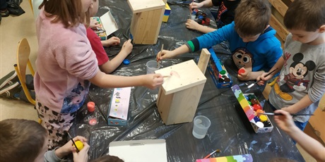 Powiększ grafikę: Uczniowie w grupie przy stolikach malują drewniane karmniki.