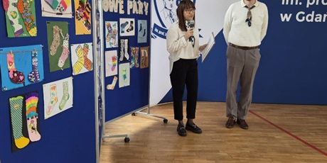 Powiększ grafikę: Piąte zdjęcie przedstawia dwóch nauczycieli, którzy mówią do mikrofonu. W tle jest widoczna tablica z pracami konkursowymi dzieci.