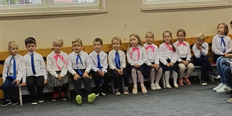 Powiększ grafikę: Uczniowie klasy pierwszej, ubrani odświętnie w białe koszule czekają na pasowanie. Dziewczyny mają różowe kokardki u szyi, a chłopcy mają niebieskie krawaty.