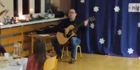 Powiększ grafikę: Szkolny nauczyciel muzyki gra na gitarze, na tle granatowej ściany. Na parapecie ozdoby świąteczne. 