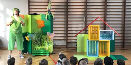 Powiększ grafikę: Aktor ubrany na zielono stoi przed dziećmi na sali gimnastycznej.