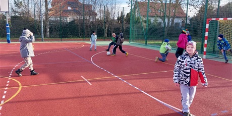 Powiększ grafikę: Dziewięcioro uczniów gra w piłkę na boisku szkolnym.