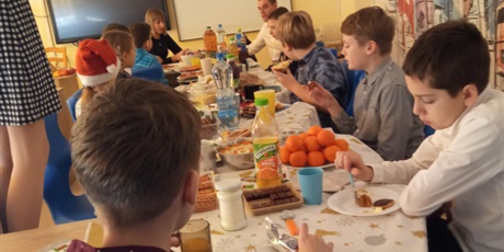 Powiększ grafikę: Uczniowie i uczennice jedzą wspólnie wigilijne przekąski przy długim stole. 
